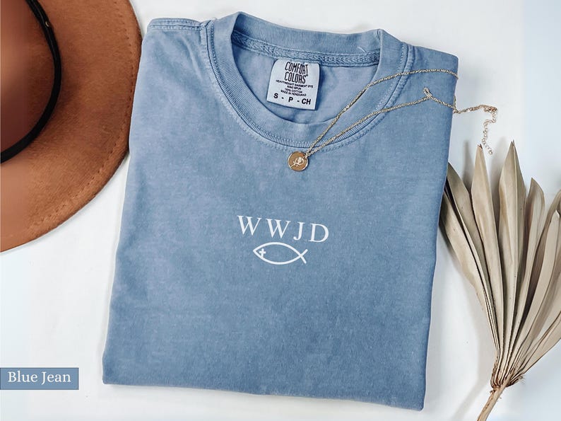 May include: A blue jean colored t-shirt with the text "WWJD" and a fish symbol in white. A gold necklace with a pendant rests on the shirt. A brown hat and dried plant are in the background.