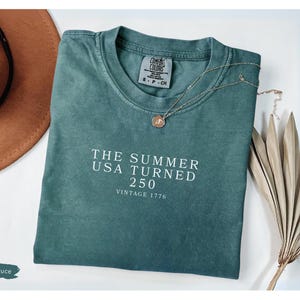 May include: Blue spruce t-shirt with the text "THE SUMMER USA TURNED 250 VINTAGE 1776" in white. A gold necklace with a pendant and a brown hat are next to the shirt. A dried plant is also in the image.