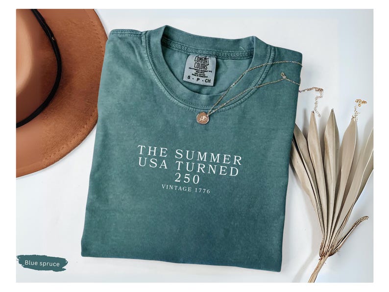 May include: Blue spruce t-shirt with the text "THE SUMMER USA TURNED 250 VINTAGE 1776" in white. A gold necklace with a pendant and a brown hat are next to the shirt. A dried plant is also in the image.