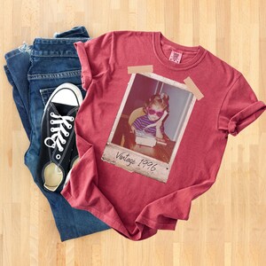 Pu&ograve; includere: Una maglietta rossa con una foto vintage di un bambino con occhiali da sole e la scritta "Vintage 1996". La maglietta &egrave; abbinata a jeans blu e scarpe da ginnastica di tela nere con lacci bianchi.