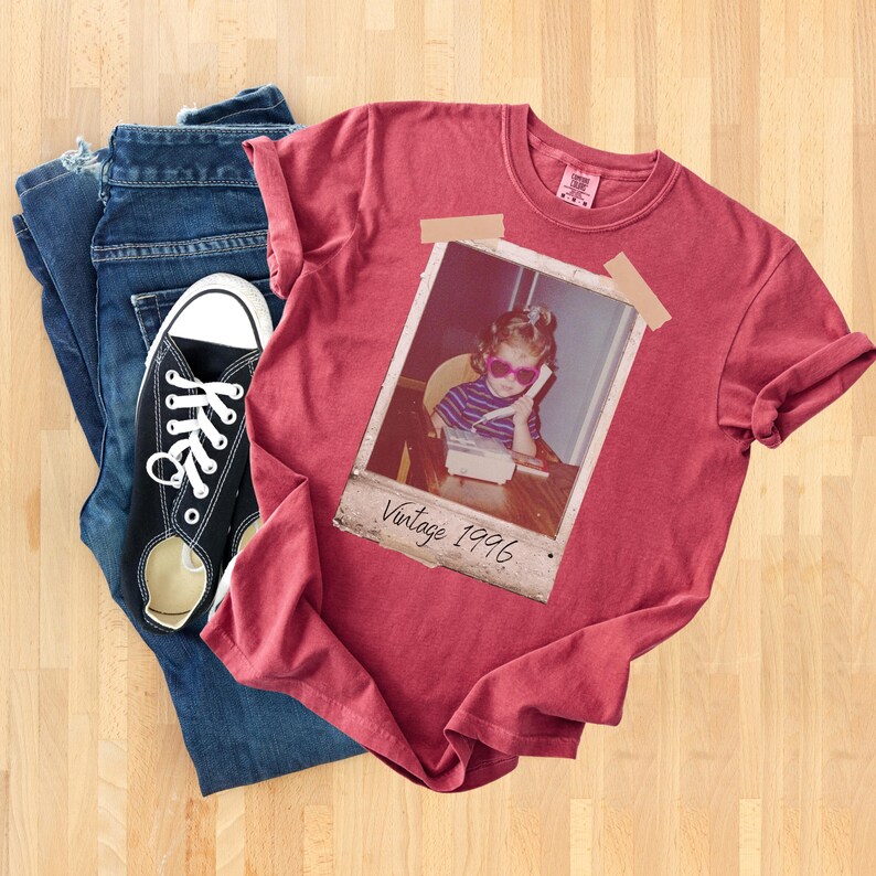 Pu&ograve; includere: Una maglietta rossa con una foto vintage di un bambino con occhiali da sole e la scritta "Vintage 1996". La maglietta &egrave; abbinata a jeans blu e scarpe da ginnastica di tela nere con lacci bianchi.