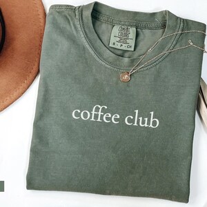 May include: A moss green t-shirt with the words "coffee club" printed in white. A gold necklace with a pendant rests on the shirt. A brown hat and dried plant are also in the image.