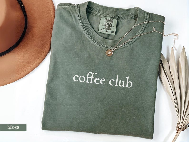 May include: A moss green t-shirt with the words "coffee club" printed in white. A gold necklace with a pendant rests on the shirt. A brown hat and dried plant are also in the image.
