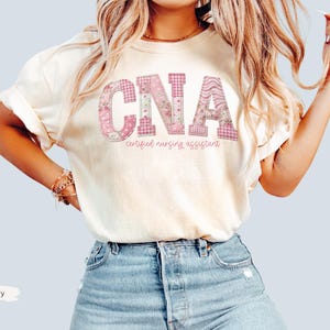May include: Ivory t-shirt featuring the letters "CNA" in a pink floral and gingham design. The words "certified nursing assistant" are printed below the letters in a cursive font. The shirt is worn with blue denim shorts.