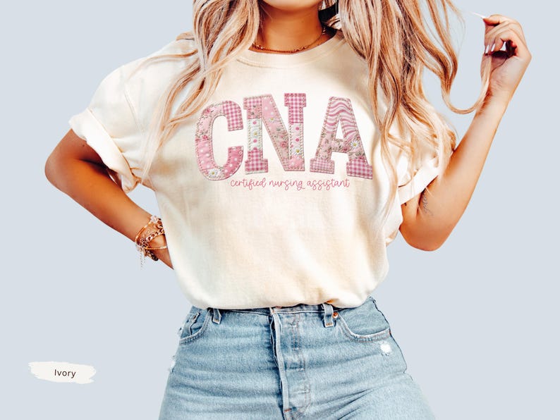 May include: Ivory t-shirt featuring the letters "CNA" in a pink floral and gingham design. The words "certified nursing assistant" are printed below the letters in a cursive font. The shirt is worn with blue denim shorts.