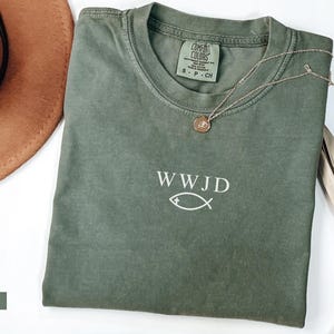 May include: A moss green Comfort Colors t-shirt with the text "WWJD" and a fish symbol. A gold necklace with a pendant rests on the shirt. A brown hat and dried plant are also in the image.