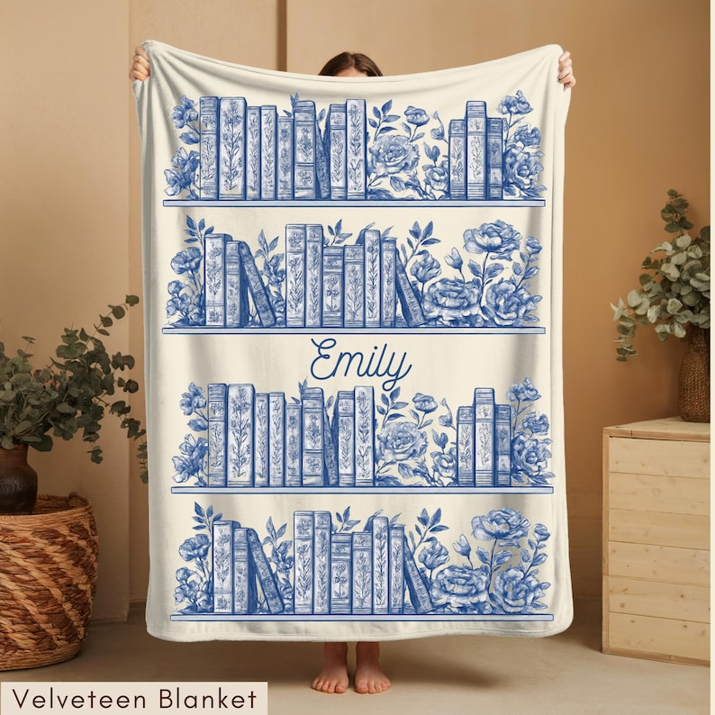 May include: Cream-colored velveeteen blanket featuring a blue bookshelf design with books, flowers, and the name "Emily." The blanket has a detailed illustration of books on shelves, with floral accents. The text "Velveteen Blanket" is at the bottom.