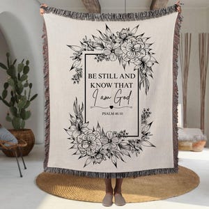 May include: Cream-colored woven throw blanket with a black floral design and the text "BE STILL AND KNOW THAT I AM GOD" from Psalm 46:10. The blanket has a fringed edge and is held up by a person.
