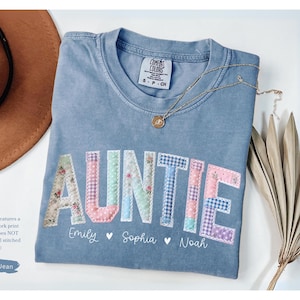 May include: A blue jean colored t-shirt with the word "AUNTIE" in a patchwork design. The shirt has the names "Emily", "Sophia", and "Noah" below the word. A brown hat, a gold necklace, and dried plants are also in the image.