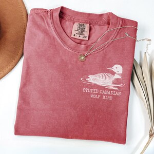 May include: Crimson-colored t-shirt with a round neck, featuring a white graphic of a loon and the text "STUPID CANADIAN WOLF BIRD." A silver necklace with a pendant is on the shirt. A brown hat and dried plant are also in the image.