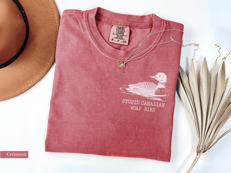 May include: Crimson-colored t-shirt with a round neck, featuring a white graphic of a loon and the text "STUPID CANADIAN WOLF BIRD." A silver necklace with a pendant is on the shirt. A brown hat and dried plant are also in the image.