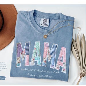 May include: A blue jean Comfort Colors t-shirt with the word "MAMA" in patchwork letters. The letters are decorated with floral and polka dot patterns. Below the word "MAMA" are names and hearts. A gold necklace and a brown hat are also in the image.