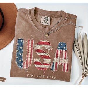 Puede incluir: Camiseta color espresso con la palabra "USA" en parches desgastados de la bandera estadounidense. La camiseta tiene cuello redondo y el texto "VINTAGE 1776" debajo. Tambi&eacute;n se ven un sombrero marr&oacute;n y un collar plateado.
