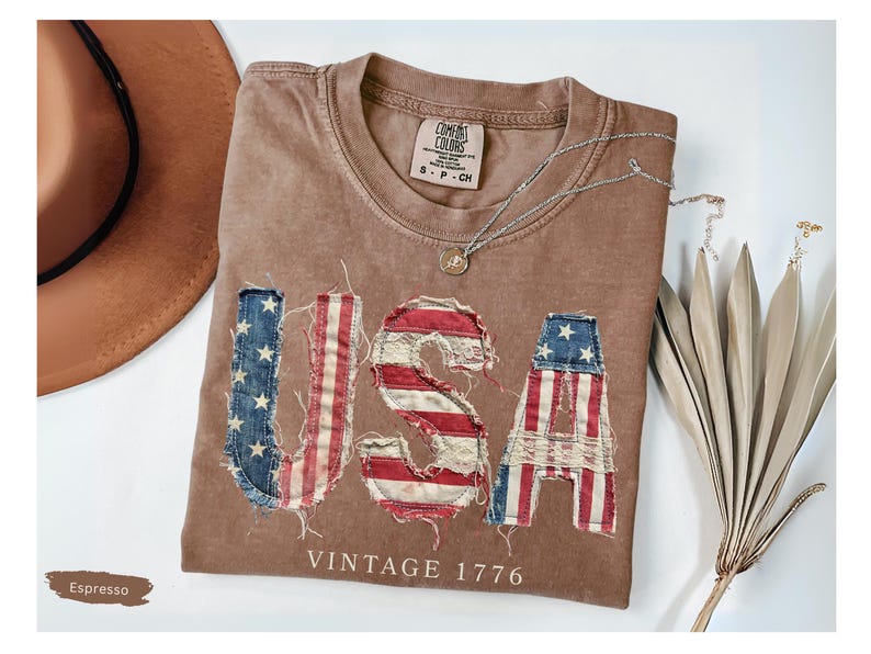 Puede incluir: Camiseta color espresso con la palabra "USA" en parches desgastados de la bandera estadounidense. La camiseta tiene cuello redondo y el texto "VINTAGE 1776" debajo. Tambi&eacute;n se ven un sombrero marr&oacute;n y un collar plateado.