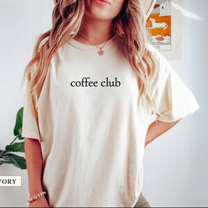 May include: Cream-coloured t-shirt featuring the words "coffee club" in black. The shirt has short sleeves and a crew neck. The text is centrally placed on the front. The material appears soft and comfortable.