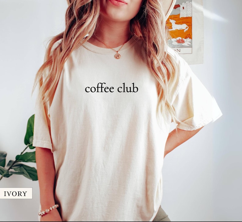 May include: Cream-coloured t-shirt featuring the words "coffee club" in black. The shirt has short sleeves and a crew neck. The text is centrally placed on the front. The material appears soft and comfortable.