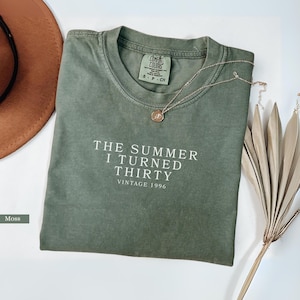 May include: A moss green t-shirt with the text "THE SUMMER I TURNED THIRTY VINTAGE 1996" in white. A gold necklace with a pendant is on the shirt. A brown hat and decorative dried plant are in the background.