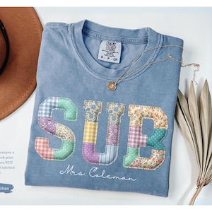 May include: Blue jean colored t-shirt with the word "SUB" in a faux patchwork design. The shirt has a crew neck and the name "Mrs. Coleman" is written in script below the letters. A gold necklace and a brown hat are also in the image.