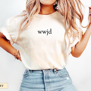 Puede incluir: Camiseta de color marfil con la inscripci&oacute;n "wwjd" en negro. La camiseta tiene un corte relajado y se combina con vaqueros azules. La imagen tambi&eacute;n muestra un collar y pulseras doradas.