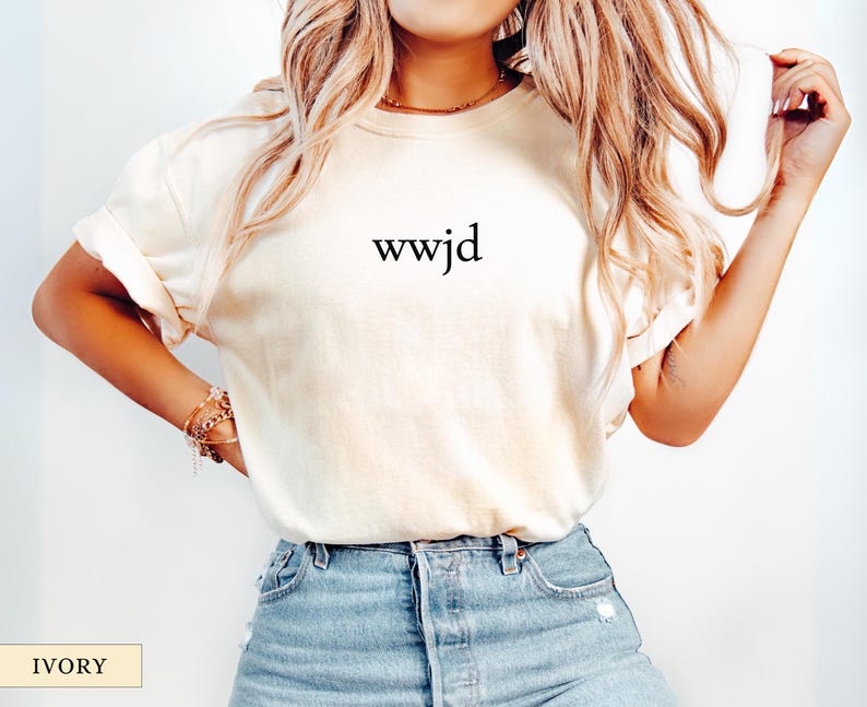 Puede incluir: Camiseta de color marfil con la inscripci&oacute;n "wwjd" en negro. La camiseta tiene un corte relajado y se combina con vaqueros azules. La imagen tambi&eacute;n muestra un collar y pulseras doradas.
