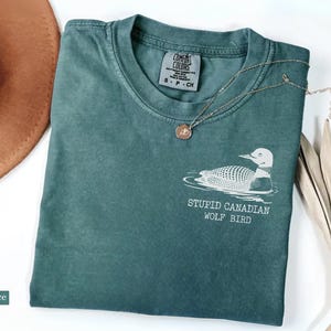 May include: Blue spruce colored t-shirt with a graphic of a loon and the text "STUPID CANADIAN WOLF BIRD". A gold necklace and a brown hat are also in the image. The shirt is folded on a white surface.