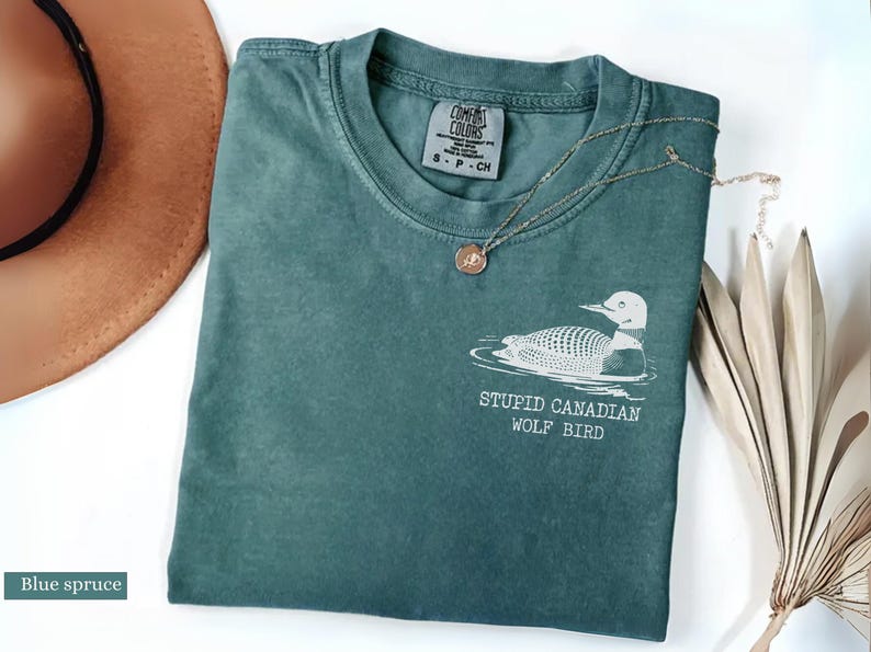 May include: Blue spruce colored t-shirt with a graphic of a loon and the text "STUPID CANADIAN WOLF BIRD". A gold necklace and a brown hat are also in the image. The shirt is folded on a white surface.