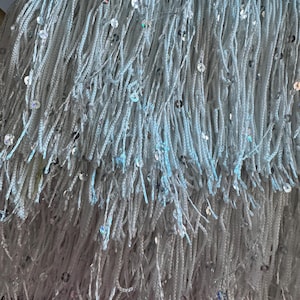 May include: Close-up of a light blue fringe trim with iridescent sequins. The fringe is made of soft, textured strands, creating a layered effect. The sequins add a touch of sparkle and glamour to the fabric.