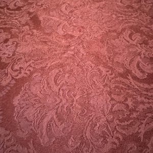 May include: Close-up of a burgundy fabric with a detailed, repeating floral pattern. The fabric has a textured appearance, with the design slightly raised from the background. The overall effect is one of elegance and sophistication, suitable for upholstery or drapery.