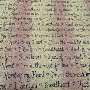 May include: A fabric with a repeating pattern of handwritten phrases in dark blue ink on a beige and pink background. Phrases include "I love you," "Sweetheart," and "Heart of my Heart."
