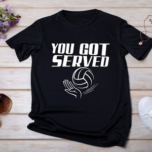 May include: Black t-shirt with white text that reads "You Got Served" and a white volleyball graphic.
