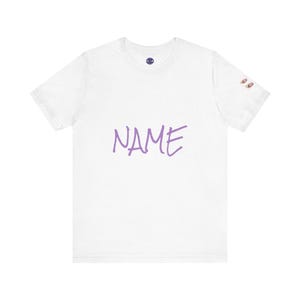May include: White t-shirt with the word "NAME" in purple script across the front. The shirt has a small graphic on the upper left chest and two small graphics on the right sleeve.