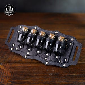 Może przedstawiać: Czarny skórzany pas na mikstury z pięcioma szklanymi fiolkami z korkowymi korkami. Pas ozdobiony jest srebrnymi ćwiekami i przeznaczony jest do przechowywania małych butelek.