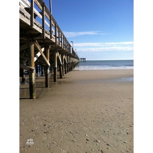 May include: A wooden pier extends out over a sandy beach with blue water in the distance. The pier is made of weathered wood and has a railing on both sides. The beach is empty and the water is calm.