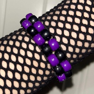 May include: A handmade bracelet featuring alternating rows of black and purple beads. The beads are cylindrical and arranged in a woven pattern. The bracelet is a vibrant accessory.