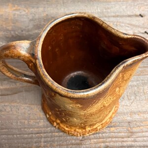 May include: A handmade ceramic pitcher with a rustic, textured appearance. The pitcher features a warm, earthy color palette with shades of brown and tan. It has a curved handle and a spout for easy pouring. The pitcher is likely used for serving beverages.
