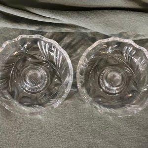 Op de afbeelding: Twee heldere glazen kommen met een bladeren ontwerp. De kommen hebben een geschulpte rand en een getextureerd midden. De kommen zijn waarschijnlijk bedoeld om te serveren of te presenteren.