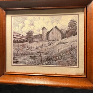 May include: A framed ink drawing on paper depicting a rural landscape with a barn, a silo, and a stream. The artwork features detailed cross-hatching and line work, set within a thick, polished medium-brown wooden frame with a beveled edge.