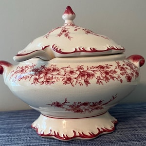 Puede incluir: Una sopera de cerámica color crema con tapa a juego y cucharón. La pieza presenta un patrón floral rojo alrededor del cuerpo y la tapa, con detalles rojos en las asas, el remate y la base festoneada. Tiene un acabado brillante y craquelado.