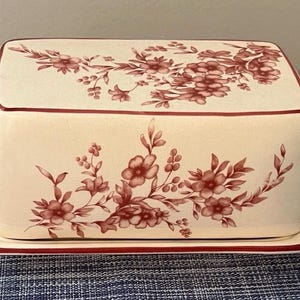 Puede incluir: Una mantequillera rectangular de cerámica con base de color crema y tapa a juego. El exterior presenta un patrón floral de color rojo oscuro con flores y hojas, con una fina línea roja oscura que bordea los bordes de la tapa y la base.