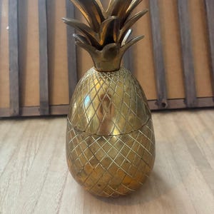 May include: A brass pineapple-shaped container with a removable lid. The body features a cross-hatched diamond pattern, and the top is adorned with pointed, overlapping leaves. The metal has a polished, golden-toned finish throughout.