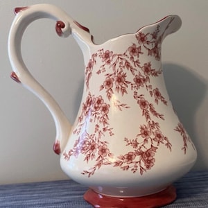 Puede incluir: Una jarra de cerámica blanca con un delicado patrón floral rojo de flores y hojas. La jarra tiene un asa curva con detalles en rojo, un pico vertedor ancho y una base roja sólida. El diseño floral envuelve el cuerpo de la pieza.