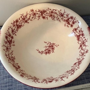Puede incluir: Un cuenco de cerámica de color crema, de forma redonda, con un patrón floral de color rojo oscuro que rodea el borde interior y un pequeño grupo floral en el centro. El cuenco incluye dos asas laterales integradas con detalles de pinceladas rojas.
