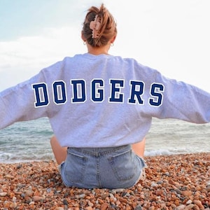 Op de afbeelding: Een lichtgrijs sweatshirt met het woord "DODGERS" in marineblauwe blokletters op de rug. Het sweatshirt heeft een witte omtrek rond de letters. De persoon zit op een kiezelstrand met de oceaan op de achtergrond.