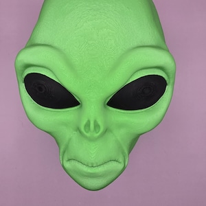 May include: A green alien mask with large black eyes and a textured surface. The mask has a smooth, rounded shape and a neutral expression. The mask is a novelty item.