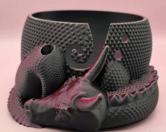 May include: A black and grey 3D printed yarn bowl shaped like a dragon. The bowl has a textured surface and a hole in the top for yarn to pass through.