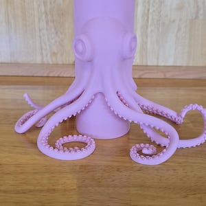 May include: A lavender octopus-shaped sculpture with a cylindrical base. The octopus has large, round eyes and multiple tentacles with small suckers. The sculpture is on a wooden surface.