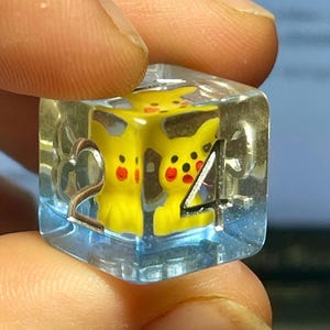 May include: A clear, blue-tinted resin die featuring two yellow Pikachu figures. The die has silver-colored metal accents and displays the numbers 2 and 4. The Pikachu figures have red cheeks and black eyes.