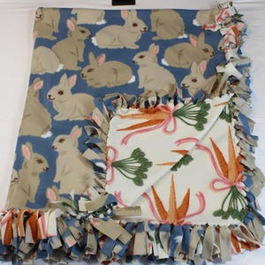 May include: A blue fleece baby blanket with a bunny rabbit pattern on one side and a carrot pattern on the other side. The blanket has a knotted fringe around the edges.