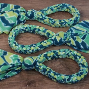 May include: Three fleece dog toys in a blue and green tie-dye pattern. Each toy has a braided loop and a knotted end with fringed fabric. The toys are arranged on a wooden surface.