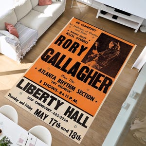 Op de afbeelding: Een rechthoekig tapijt met een oranje en crèmekleurig kleurenschema. Het tapijt bevat de tekst "RORY GALLAGHER" in grote zwarte letters, samen met concertdetails, waaronder de locatie "LIBERTY HALL" en de data "17 en 18 mei".